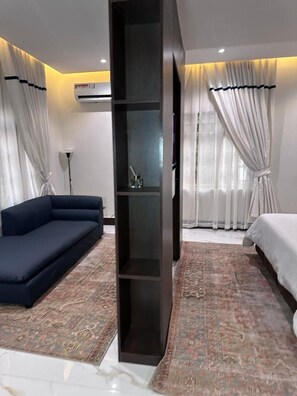 A modern hotel room with a sofa, a bed, a large rug, and a tall, dark-colored shelving unit. - Centurion Apartments - Jabi (Abuja)