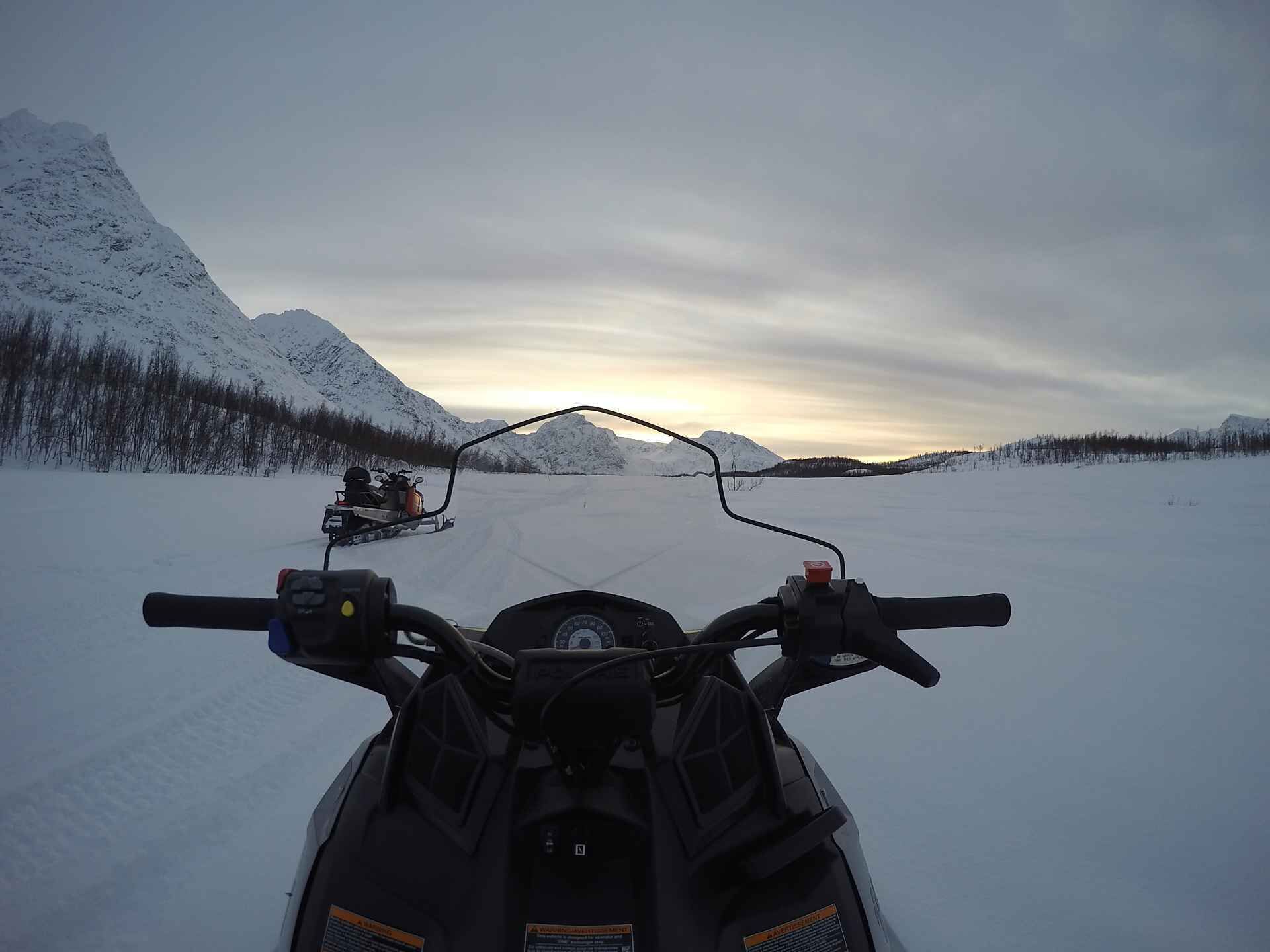 Una moto de nieve en un camino despejado, con montañas nevadas y un cielo nublado.
