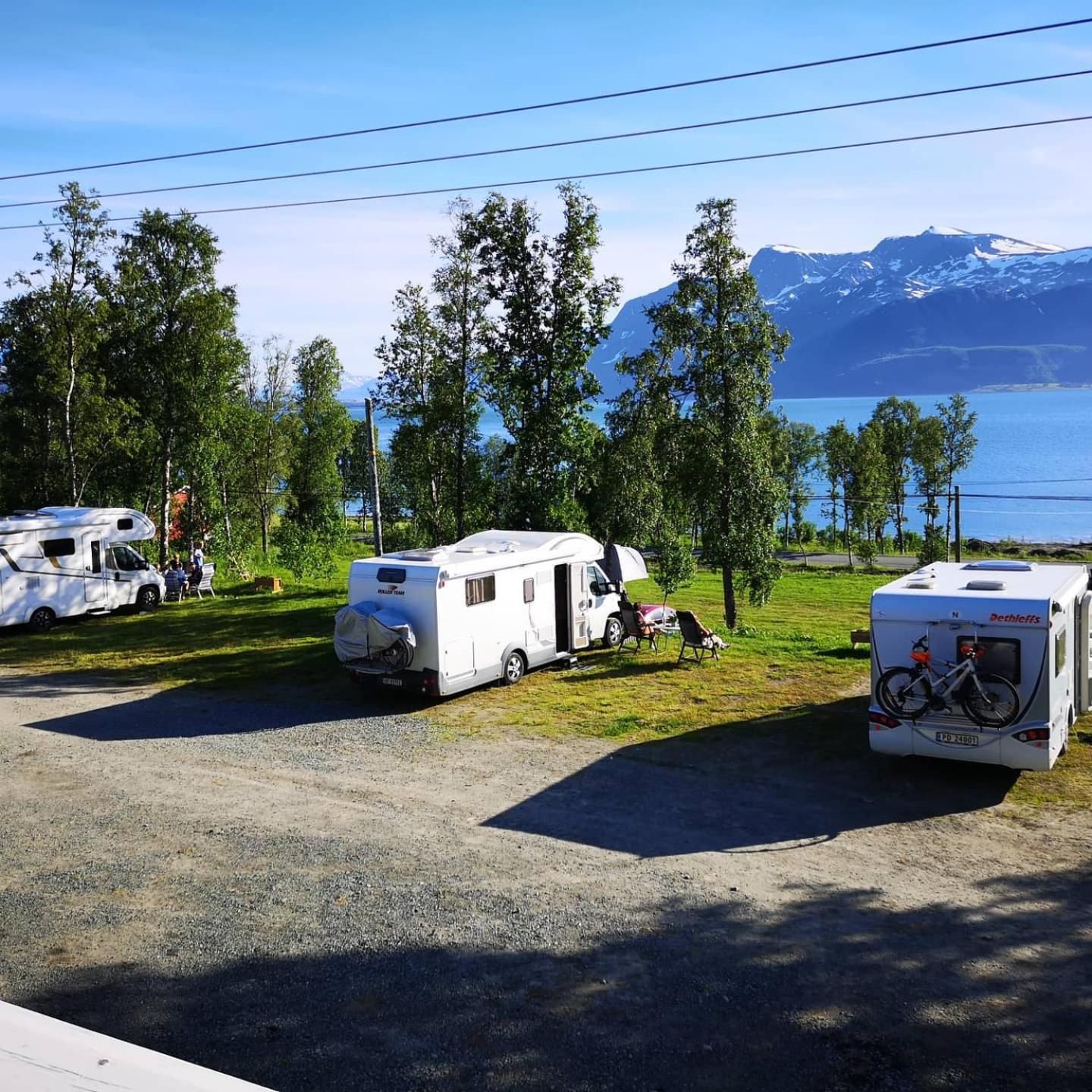 A campground with RVs parked on a gravel area, surrounded by trees and a lake with mountains in the background.