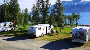Ein Campingplatz mit Reisemobilen auf Kiesflächen, umgeben von Bäumen und einem See mit Bergen im Hintergrund.