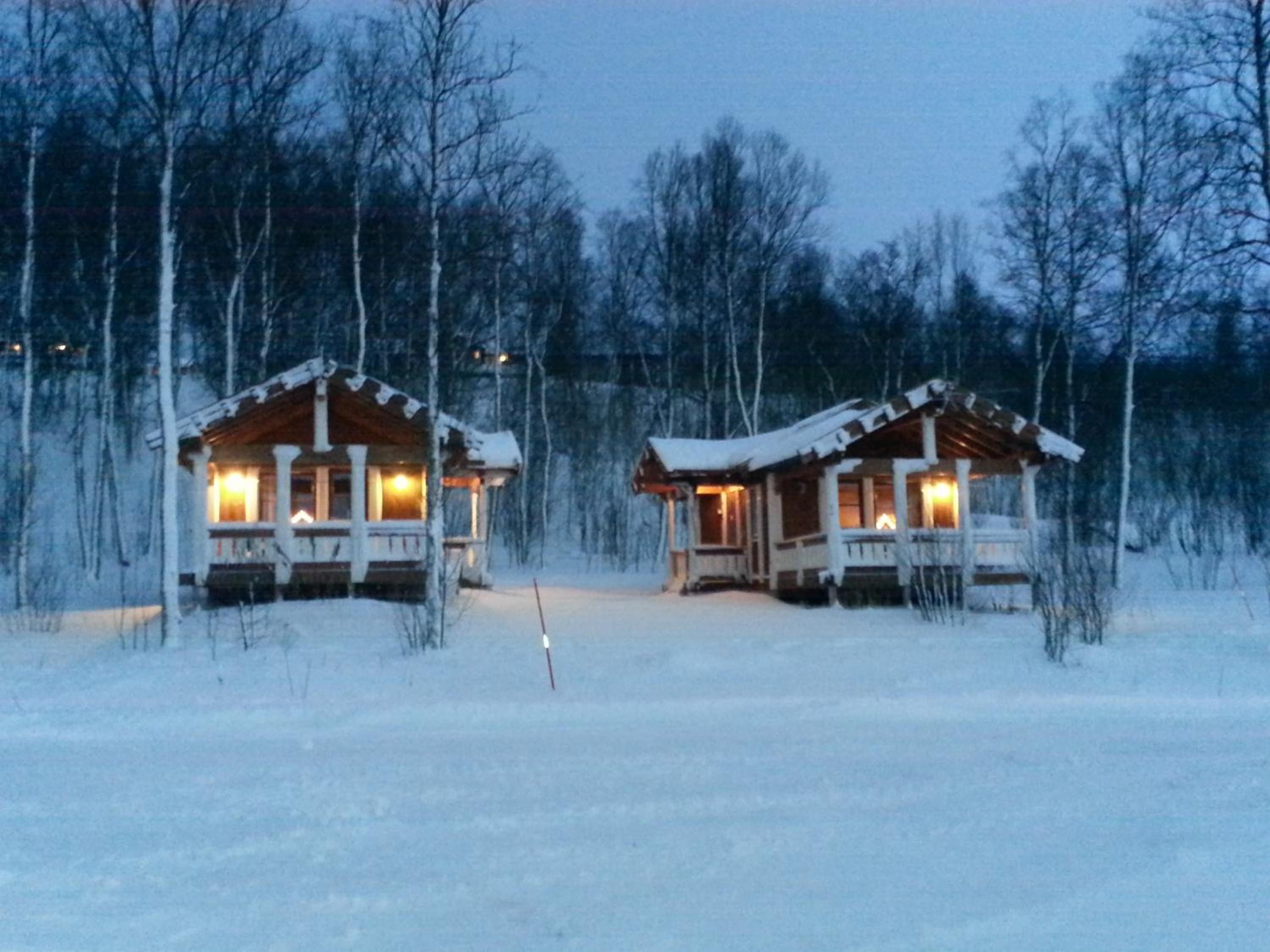 Två trähus med upplysta fönster, omgivna av snötäckta träd.
