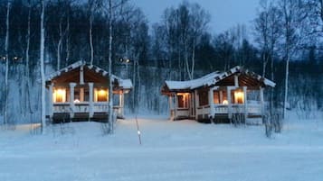 Zwei Holzhütten mit erleuchteten Fenstern, umgeben von schneebedeckten Bäumen.