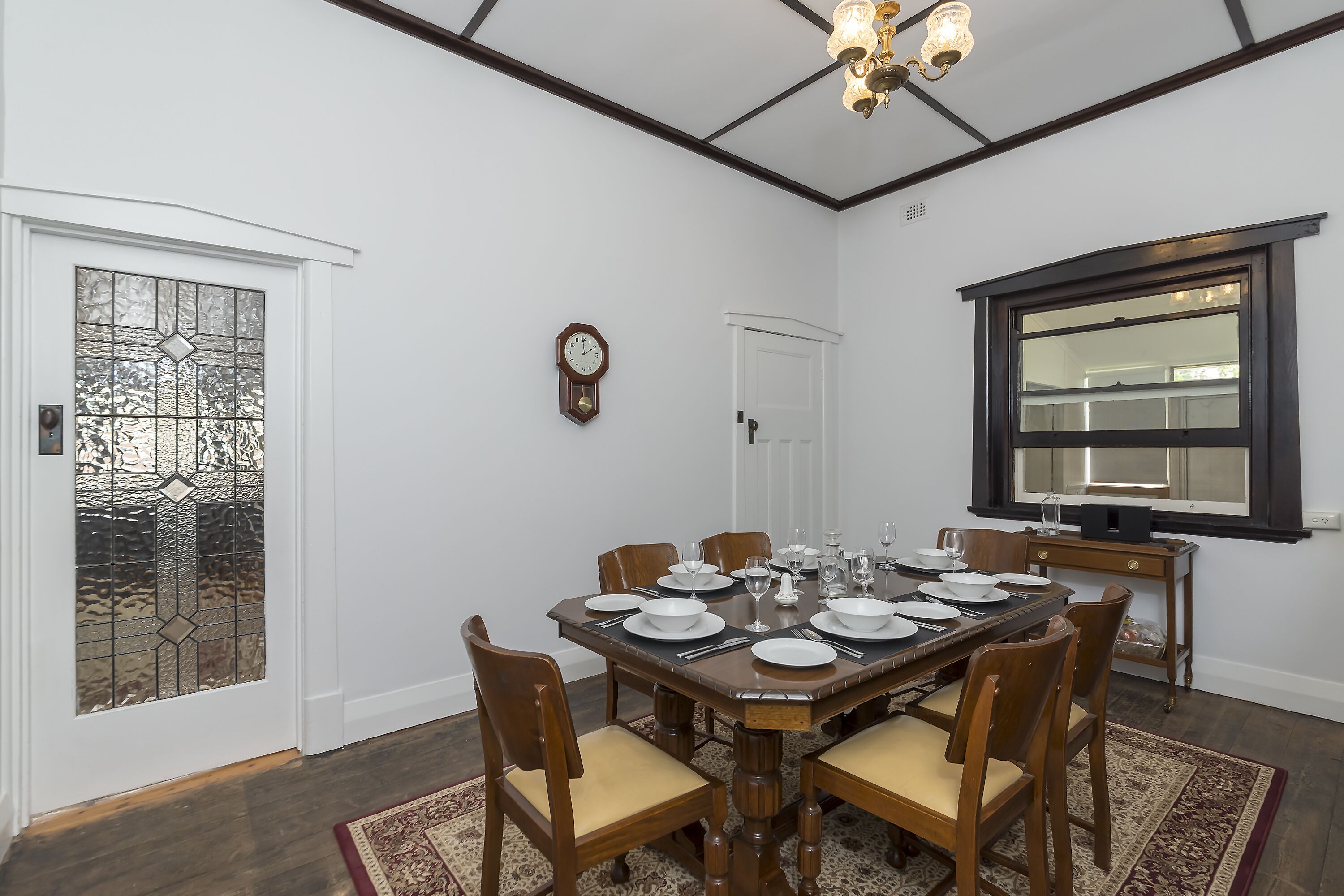 A dining room with a wooden table set for six, surrounded by chairs. A clock and a window with a dark frame are visible on the walls.