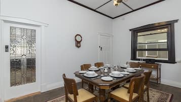 A dining room with a wooden table set for six, surrounded by chairs. A clock and a window with a dark frame are visible on the walls.