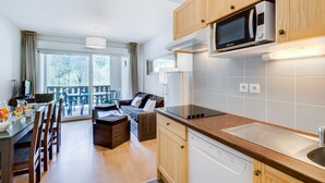 Una cocina compacta con armarios de madera, un microondas y un lavavajillas. Hay un comedor con una mesa y sillas, y un sofá al fondo.