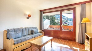 A room with a wooden sofa, a wooden coffee table, a large sliding glass door with a view of a mountainous landscape, and a small table with a lamp.