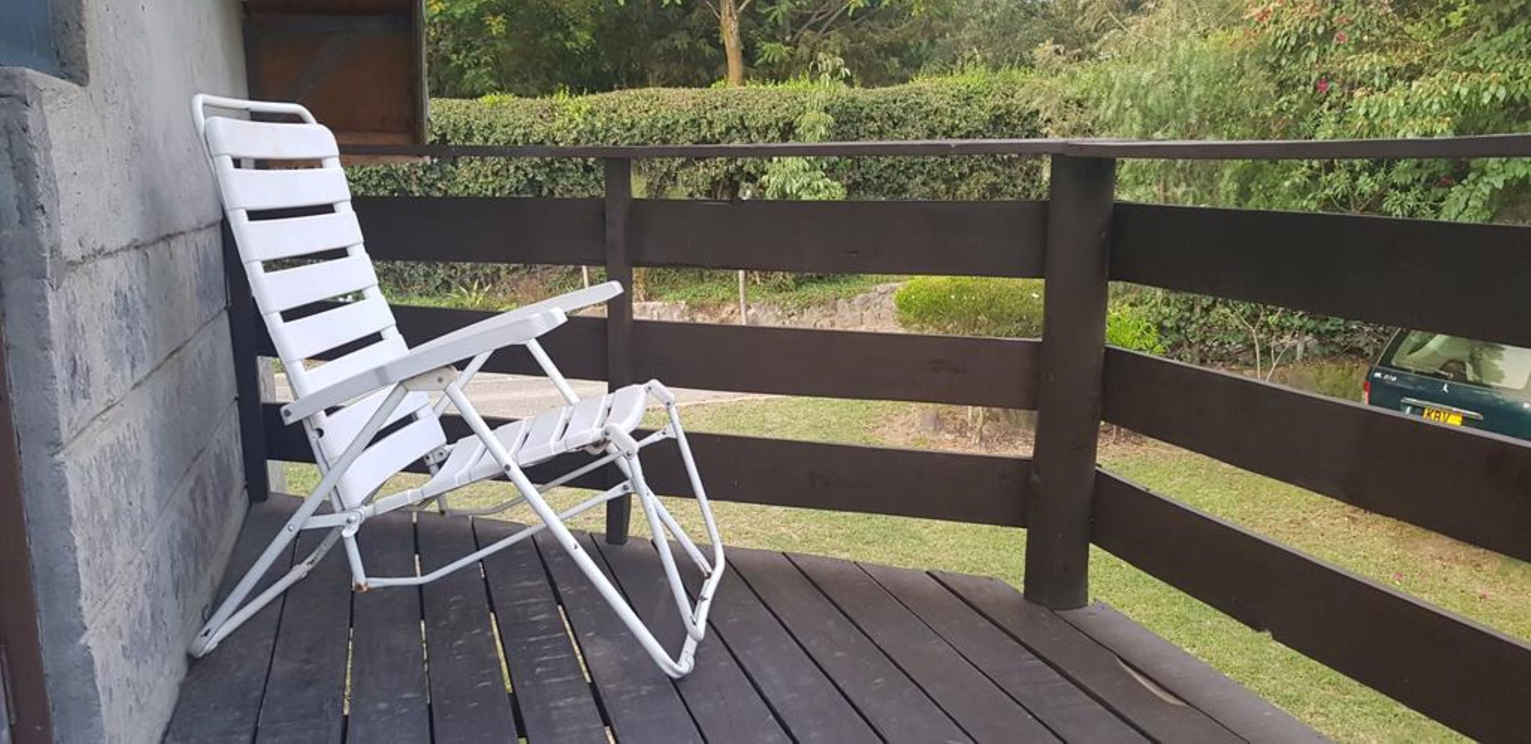 A white folding chair on a wooden deck with a view of a garden and a parked car.