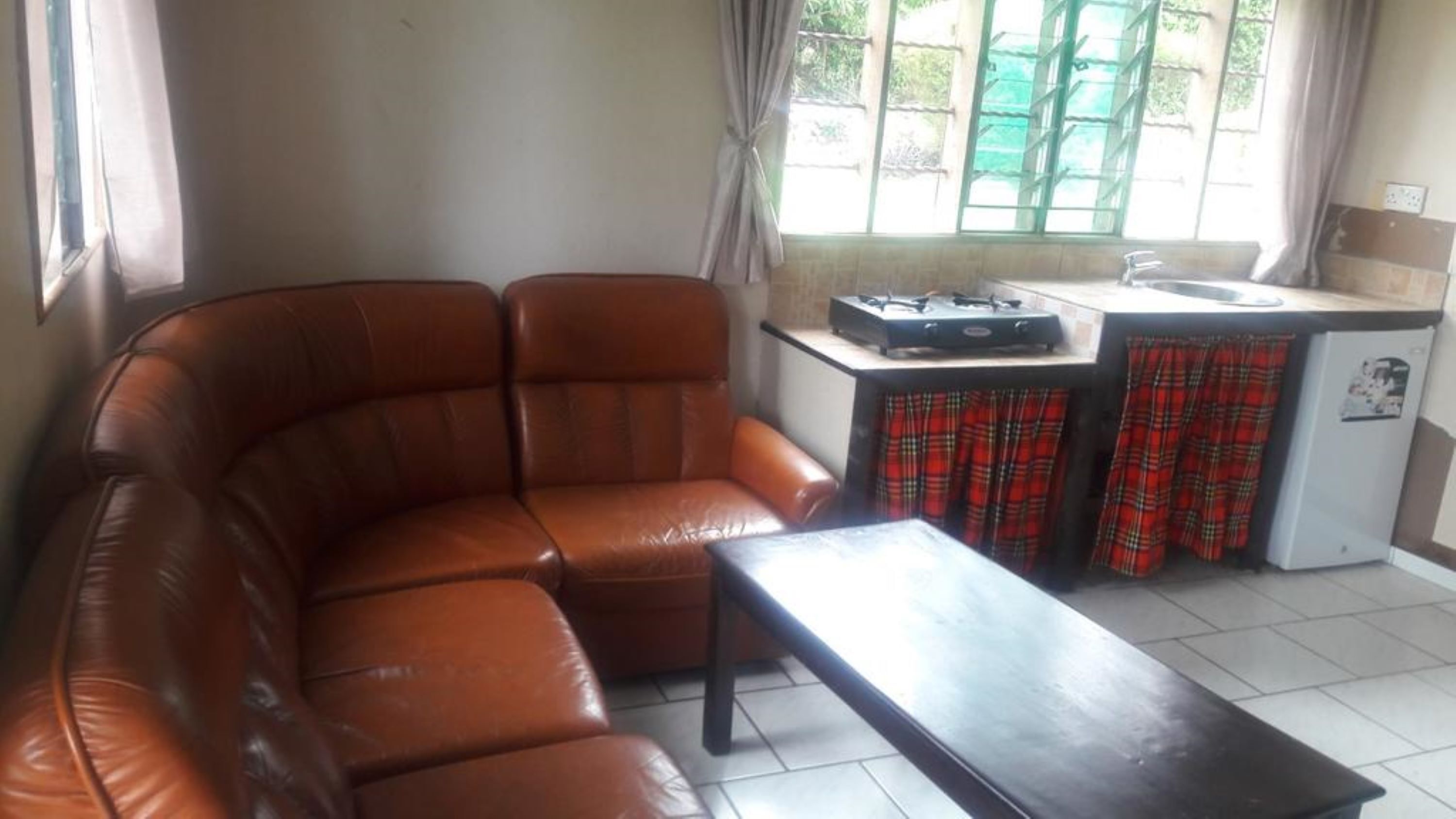 A living room with a brown leather sofa, a small wooden table, and a kitchen area with a gas stove and a refrigerator.