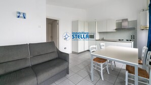 A modern living room with a grey sofa, a white dining table with four chairs, and a kitchen area with white cabinets and a stove. - Milan's Apartment n.1 (Jesolo)