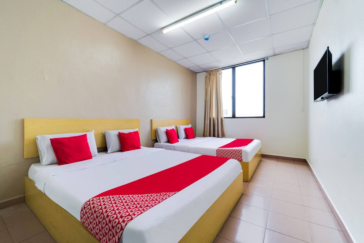 A hotel room with two beds, each with white bedding and red accents, a flat-screen TV mounted on the wall, and a window with curtains.