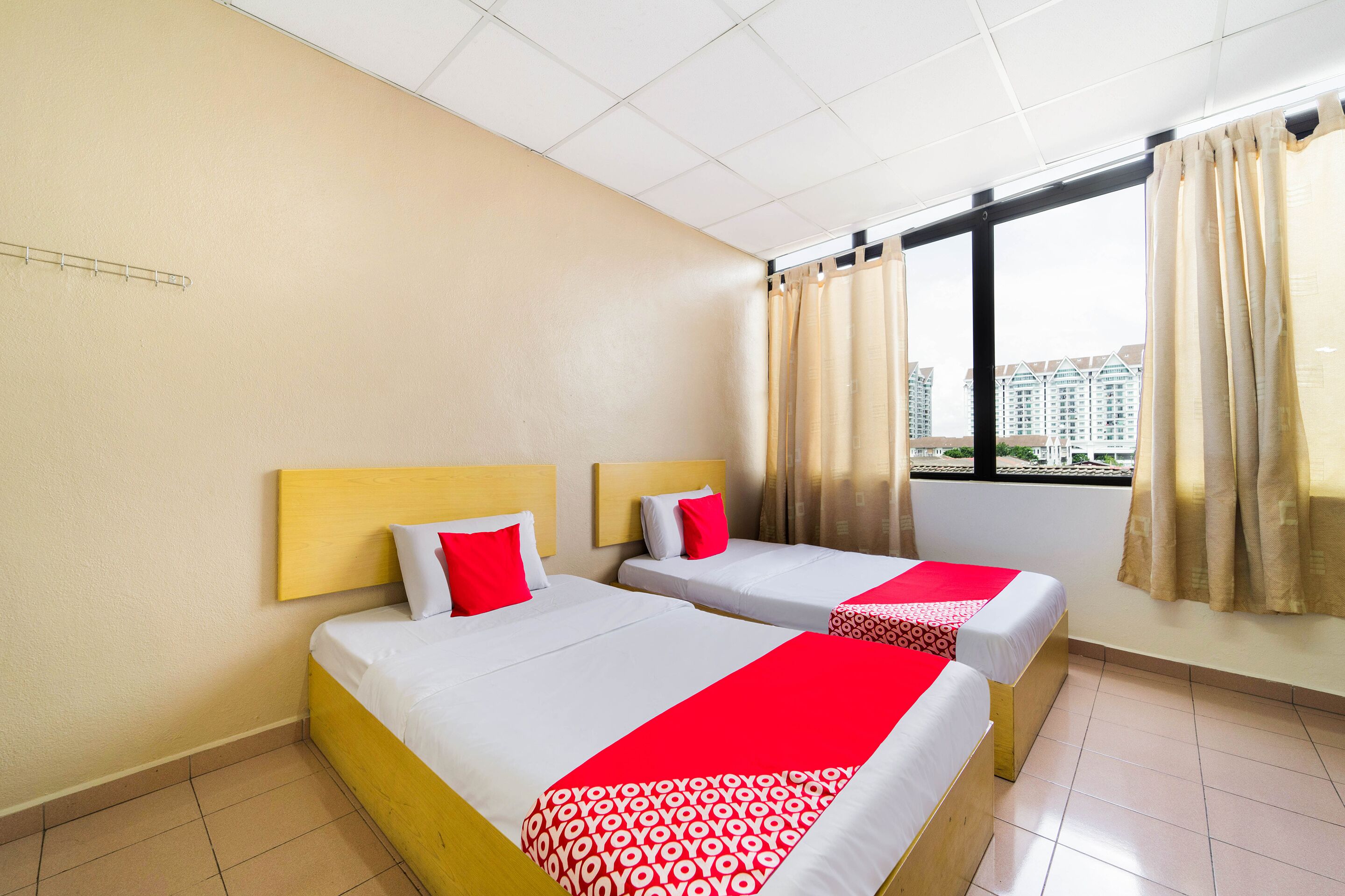 A hotel room with two beds, beige walls, a window with curtains, and a view of buildings.