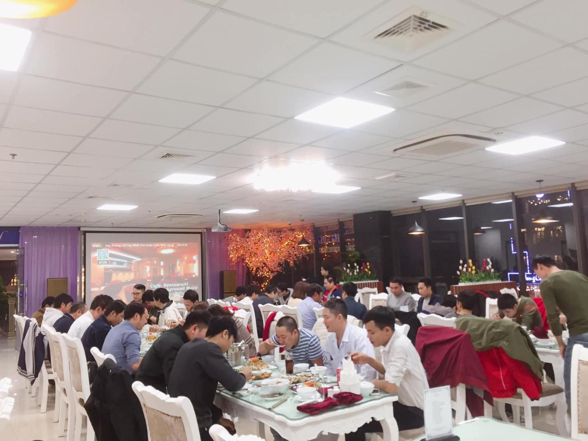 A crowded restaurant with white tables and chairs, a large screen displaying a QR code, and a decorative floral arrangement in the background.