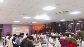 A crowded restaurant with white tables and chairs, a large screen displaying a QR code, and a decorative floral arrangement in the background.