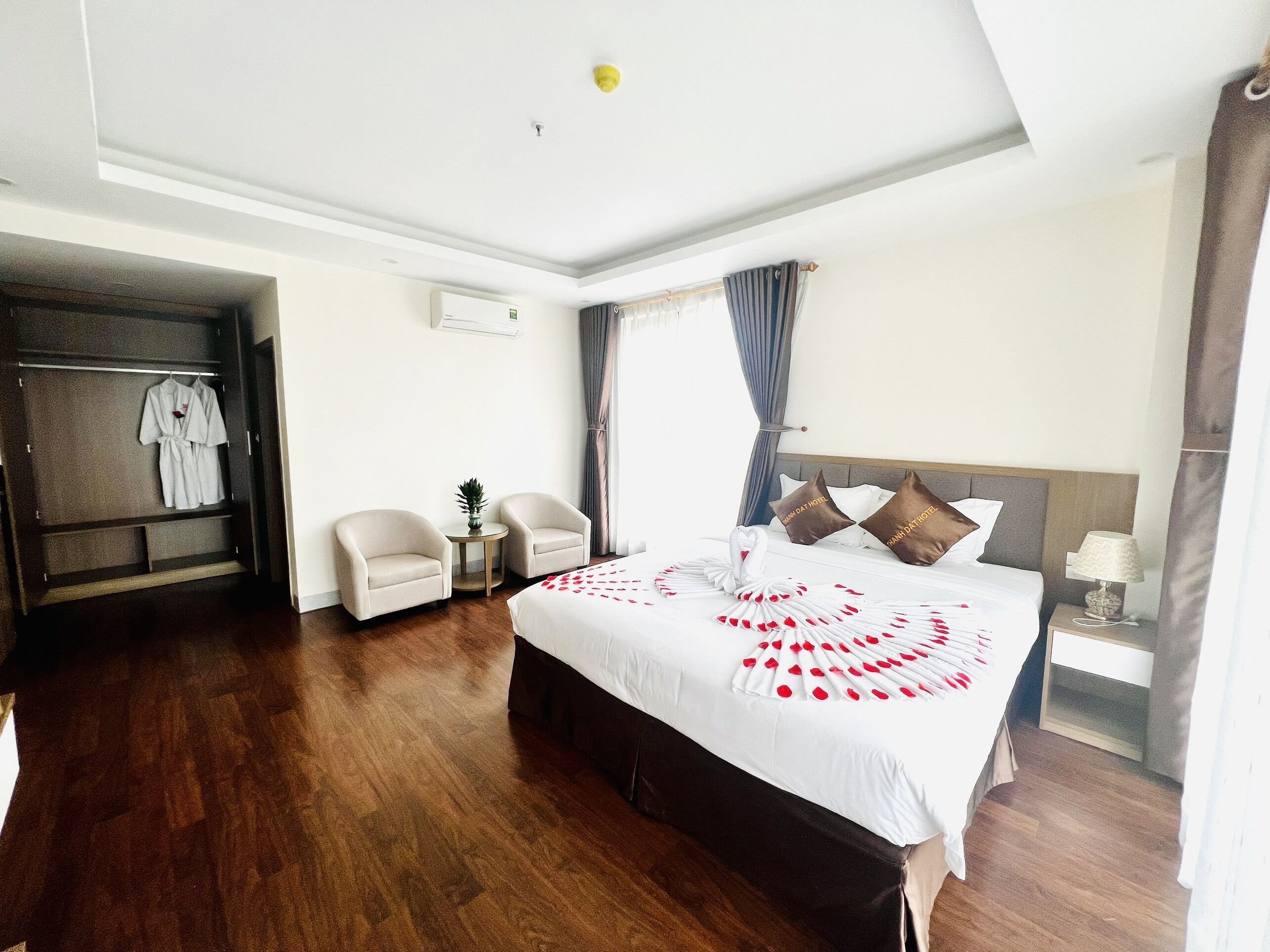 A hotel room with a large bed, a wardrobe, two armchairs, a small table with a plant, and a nightstand with a lamp.