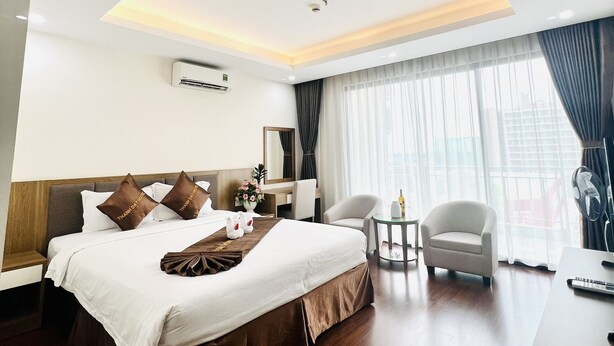 Ein modernes Hotelzimmer mit einem großen Bett, einer Sitzecke mit Sessel und Tisch sowie einem großen Fenster mit Vorhängen.