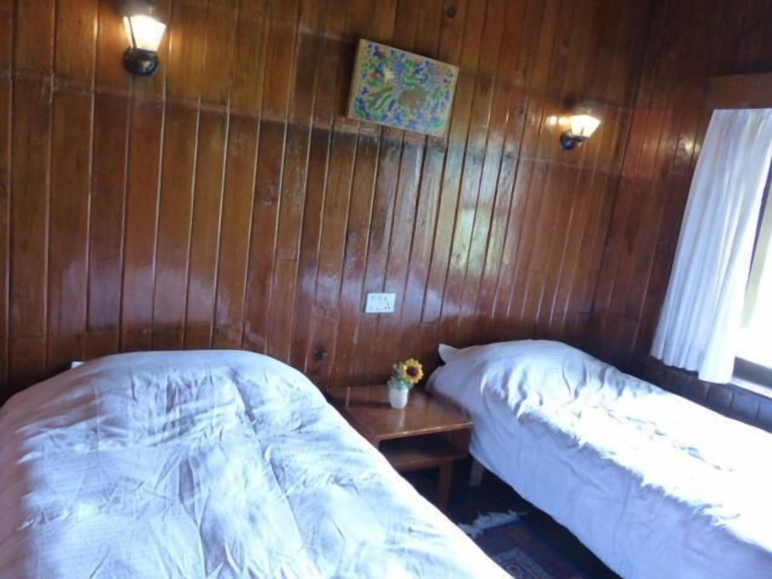 A room with wooden panel walls, two beds with white bedding, a small table with a vase of flowers, and a painting on the wall.