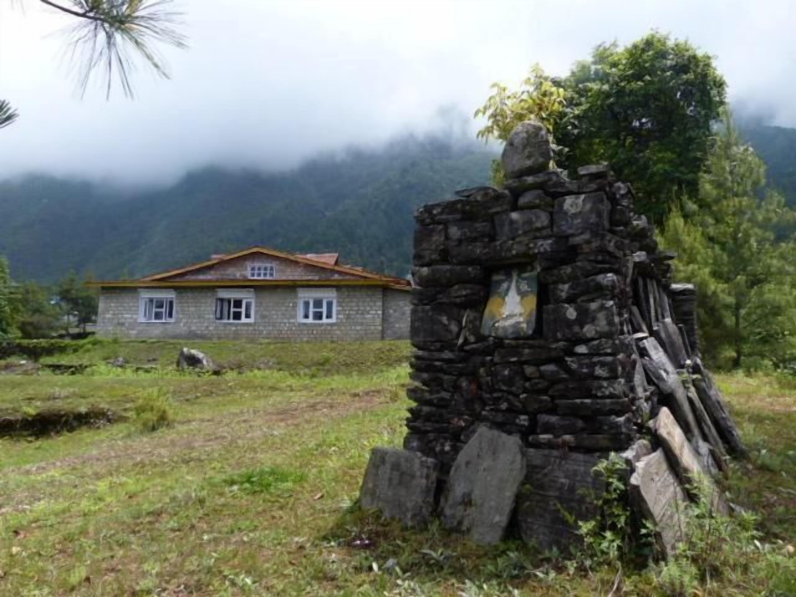 A stone structure with a window, a stone wall, and a house with a red roof in a grassy area with trees and mountains in the background.