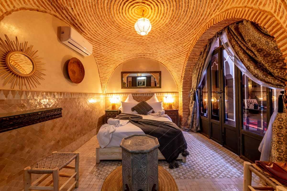 A room with a patterned ceiling, a bed with pillows, a nightstand, a small table, and a mirror on the wall.
