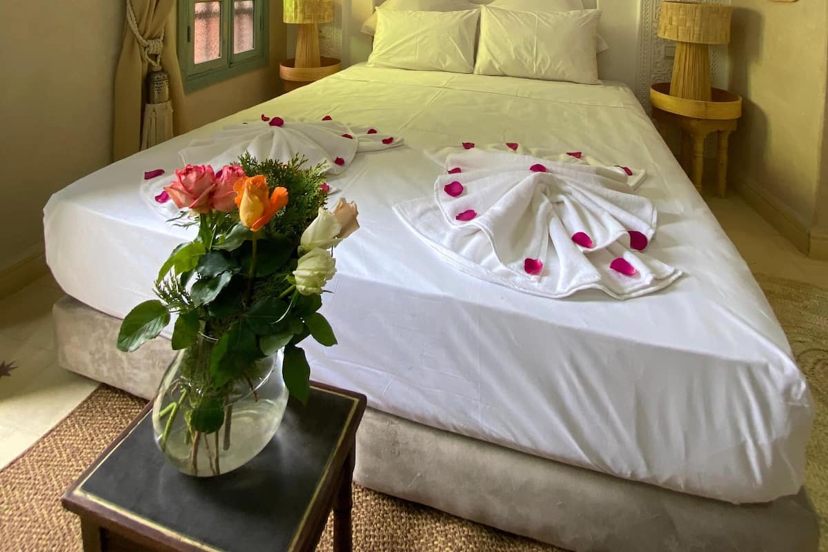 A neatly made bed with white linens and rose petals, a vase with flowers on a bedside table, and a decorative mirror on the wall.