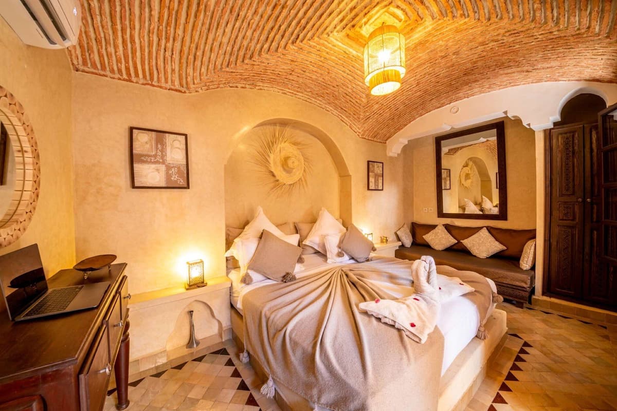 A spacious bedroom with a large bed, a desk with a laptop, a mirror, and a decorative ceiling.