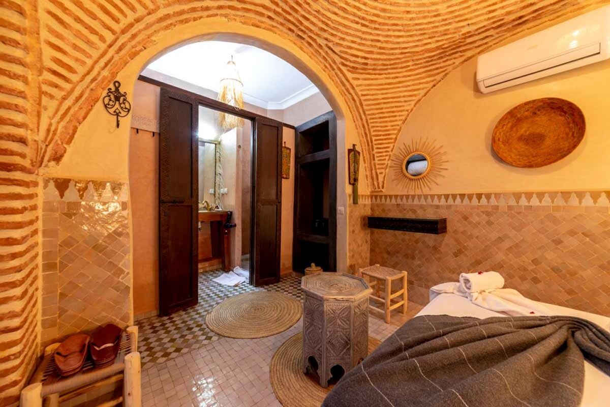 Hotelkamer met een zwart-wit geruit vloertje, een bed met dekbed, een klein rond tafeltje, een stoel, een handdoek, en een boogvormige deuropening naar een badkamer.