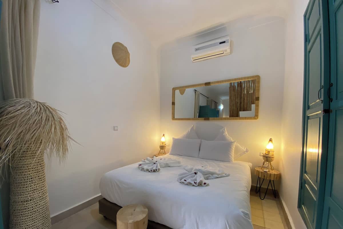 A neatly made bed with white linens, a round wooden stool, a woven basket, and a wall-mounted air conditioning unit.