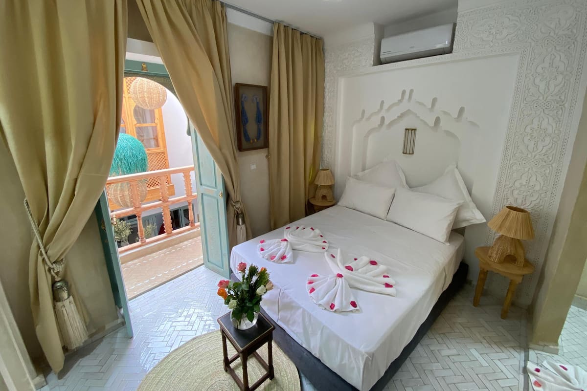 A hotel room with a bed, a nightstand, a lamp, a small table with a vase of flowers, and a view of an outdoor area through a door.