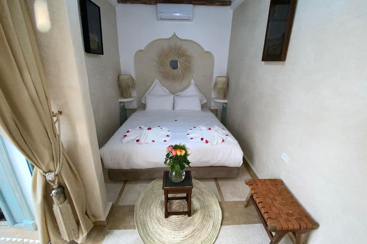 A bedroom with a bed, a small table with a vase of flowers, a bench, and a wall-mounted air conditioning unit.