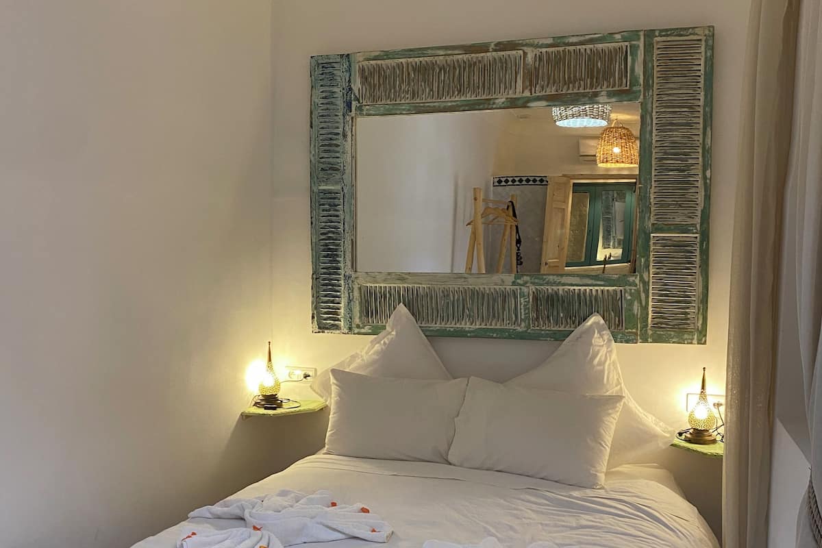 A neatly made bed with white linens and towels, a large mirror with a distressed frame, and two bedside lamps on wooden nightstands.