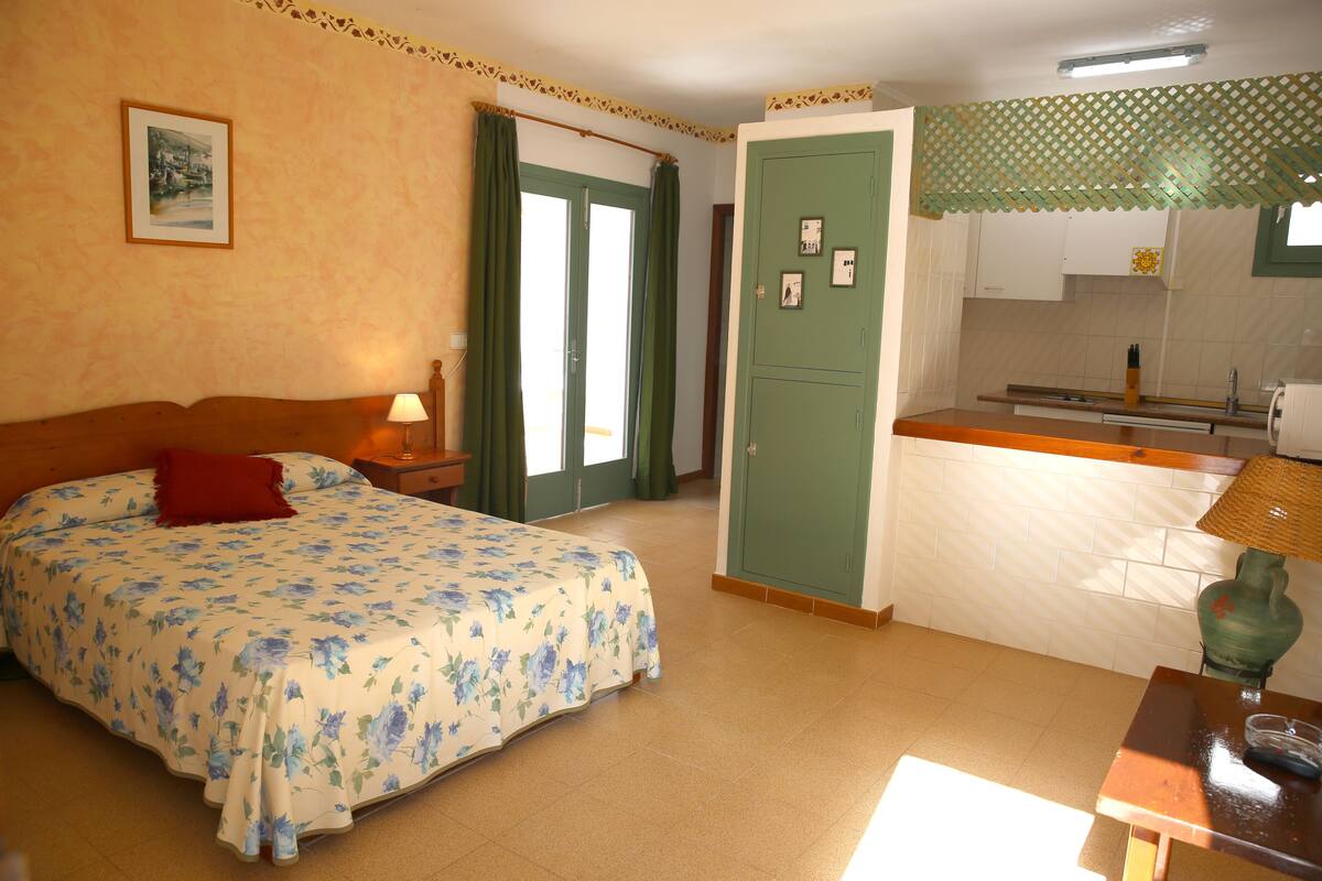 A bedroom with a bed, a nightstand, a lamp, a green wardrobe, and a kitchenette with a sink and countertop.