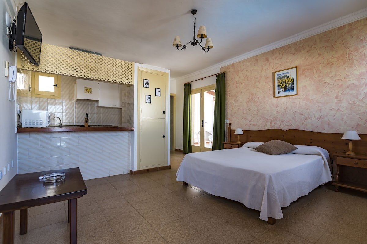 A compact hotel room with a kitchenette, a bed with a wooden headboard, a small table, and a nightstand with a lamp.