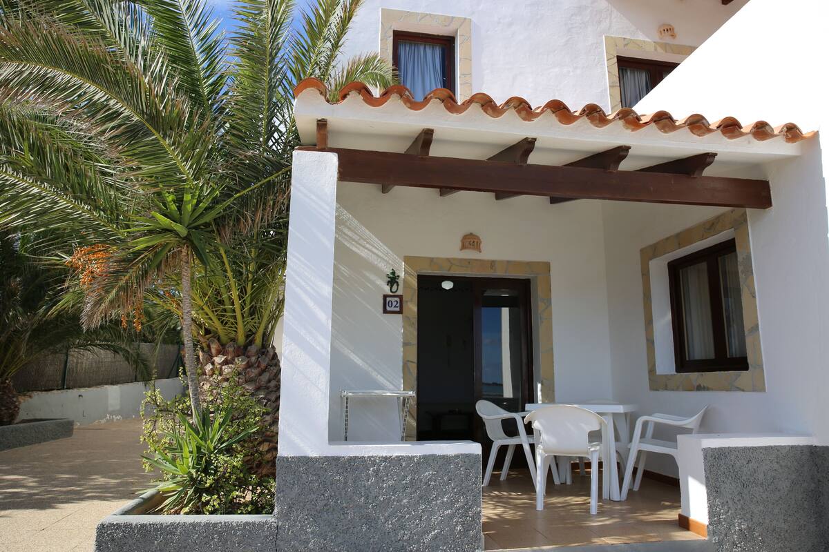 A covered outdoor seating area with white plastic chairs and a table, a door with a number sign, and a palm tree.