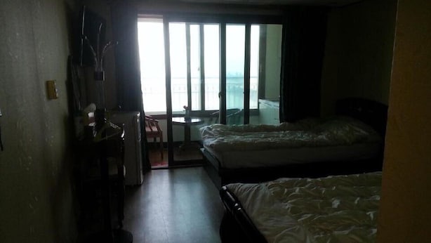 A hotel room with two beds, a small table, and a view of the sea through large windows. - Eden Hostel (Jeju City)