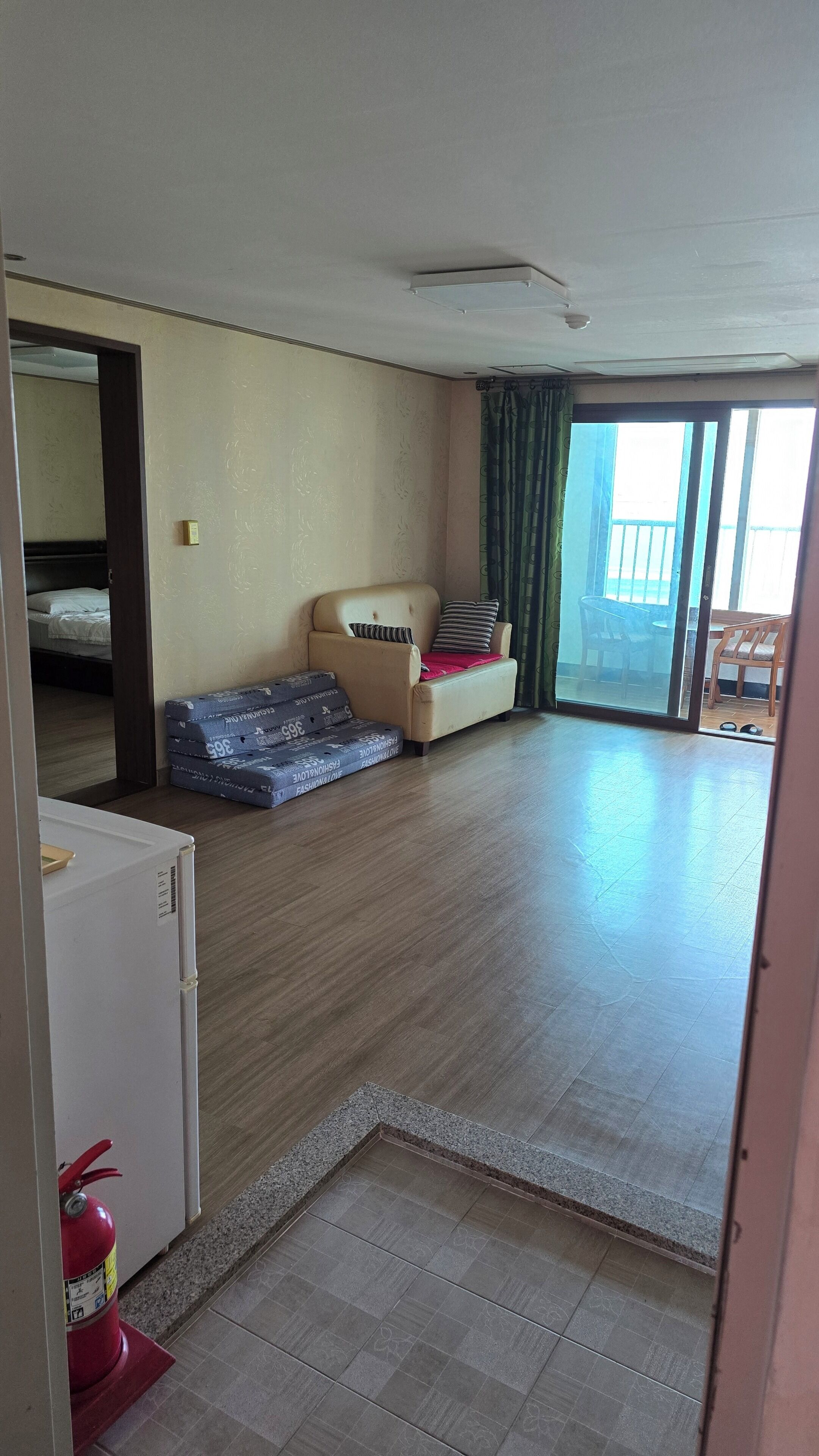 A room with a sofa, a small table, a fire extinguisher, and a sliding glass door leading to an outdoor area.