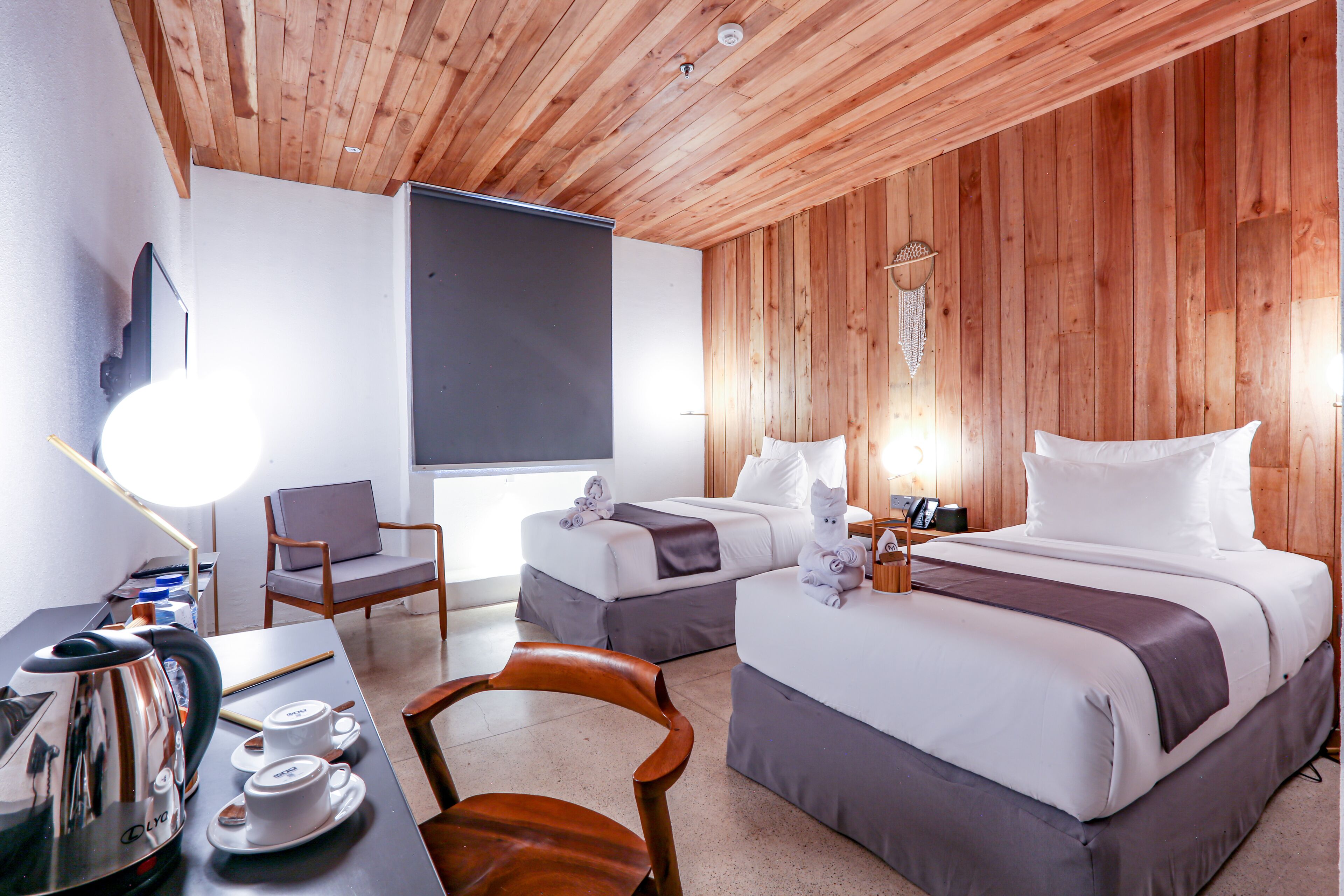 A hotel room with two beds, a desk with a kettle and cups, a chair, and a wooden ceiling.