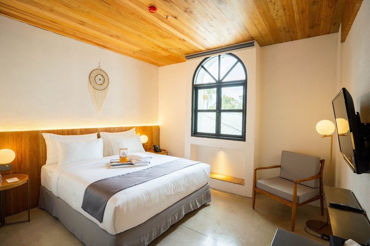 A hotel room with a large bed, wooden ceiling, a window with a view, a desk with a chair, and a wall-mounted TV.