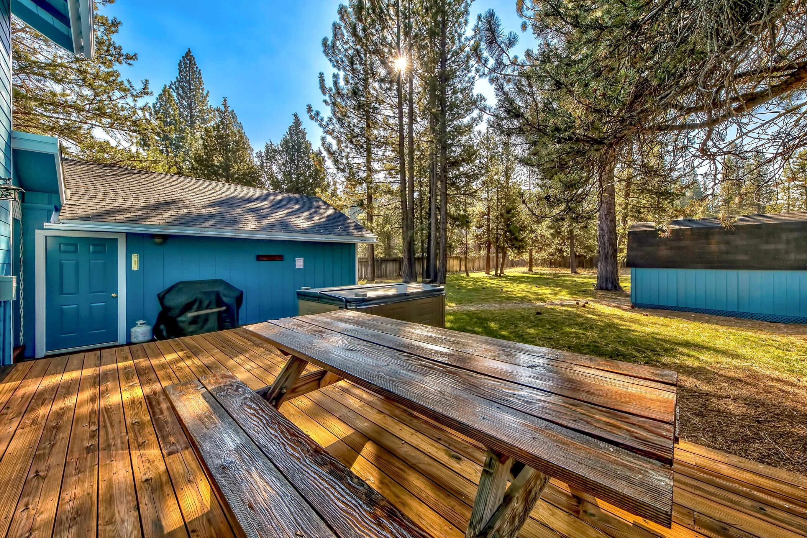 Une terrasse en bois avec une table de pique-nique, un bâtiment bleu avec un garage, et une zone boisée.