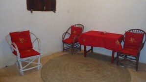 A room with a round woven rug, two white metal chairs, and a wooden table with a red tablecloth. - Dudu Villas n Cottages (Lamu)