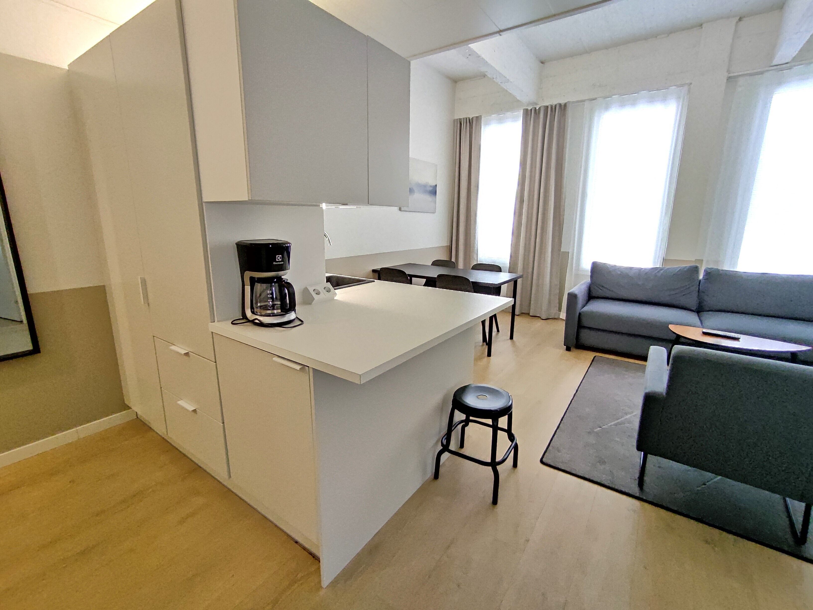 A modern kitchen with a coffee maker, a dining area with a sofa and a small table, and a large window with curtains.