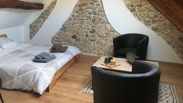 A bedroom with a stone wall, a bed with a white and gray patterned bedspread, a black chair, and a small wooden table with a cup and saucer. - Les chambres d'Orsay (Orsay)