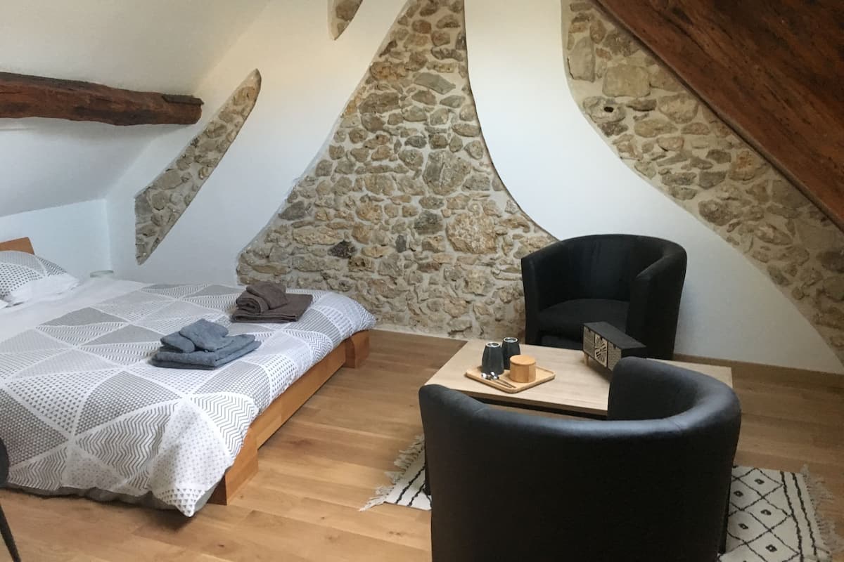 Une chambre avec un mur en pierre, un lit recouvert d’une courtepointe à motifs blancs et gris, une chaise noire et une petite table en bois sur laquelle se trouvent une tasse et une soucoupe.