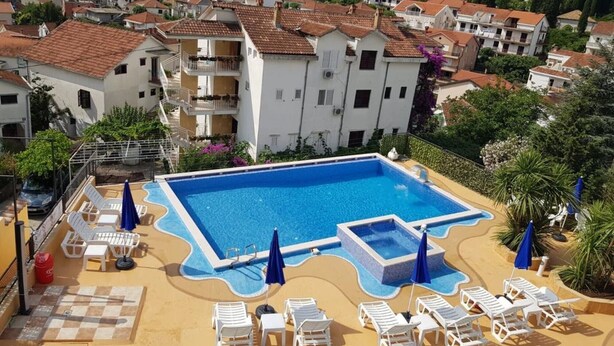 Piscina com espreguiçadeiras e guarda-sóis, cercada por edifícios com telhados de telha vermelha.