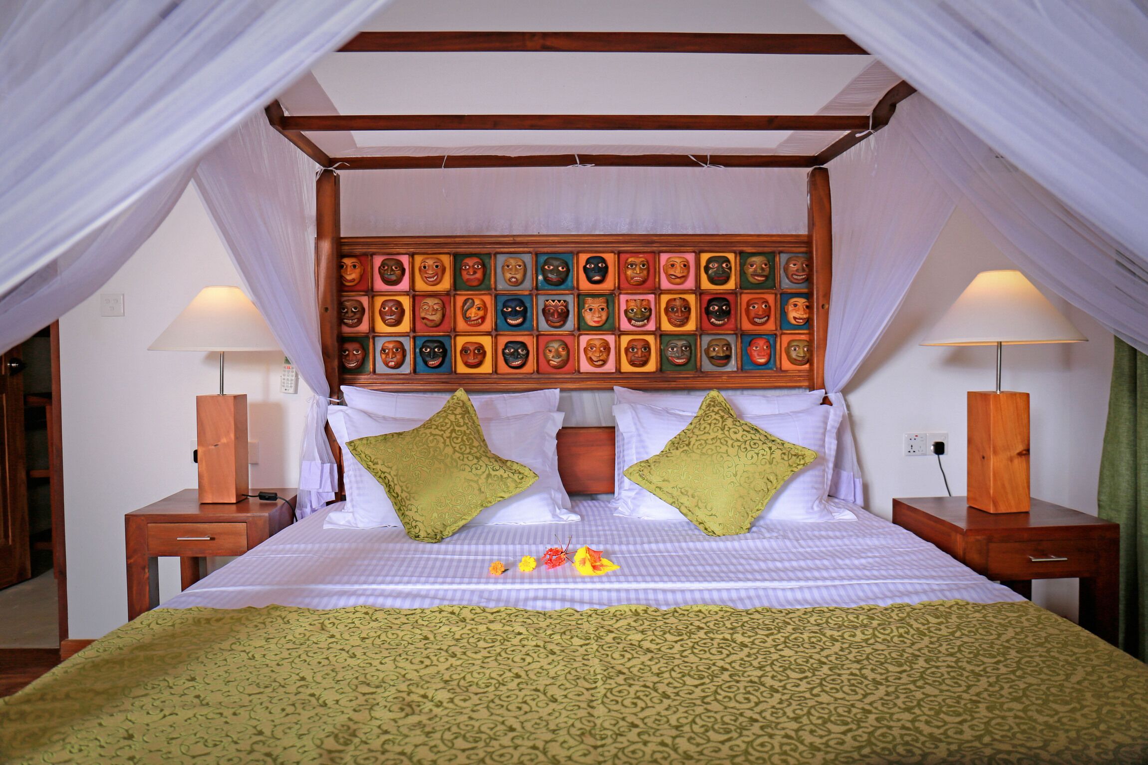 Un lit à baldaquin, avec des colonnes aux quatre coins, une tête de lit ornée de masques colorés, des coussins verts et une couvre-lit à motifs jaunes.