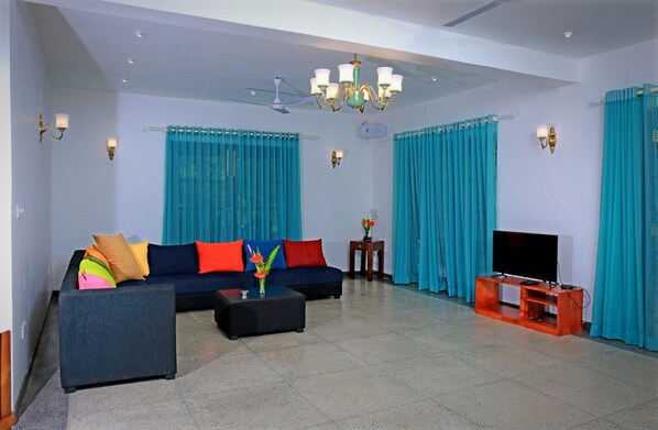 A living room with a dark blue sofa, colorful cushions, a coffee table, a TV on a wooden stand, and teal curtains.