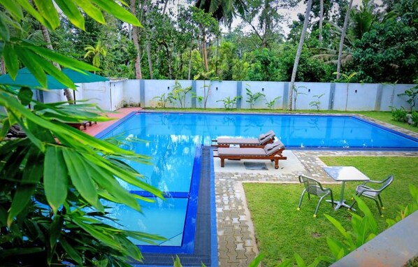 A rectangular swimming pool with a blue interior, surrounded by a white wall and lush greenery. There are two lounge chairs and a small table on the poolside.