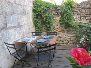 Una mesa de madera dispuesta con platos, cubiertos y vasos, rodeada de sillas de metal, frente a una pared de piedra con plantas.