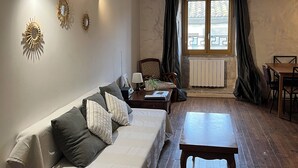 Un salon chaleureux avec un plancher en bois, un canapé, une table basse et une table à manger. Il y a une fenêtre avec des rideaux, un radiateur et des lampes fixées au mur.