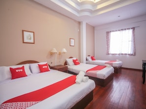 A hotel room with three beds, each with red and white bedding and a red 'OYO' pillow. There are two wall-mounted lamps, a window with floral curtains, and a wooden floor. - OYO 89537 Fairy Garden Resort (Ranau)