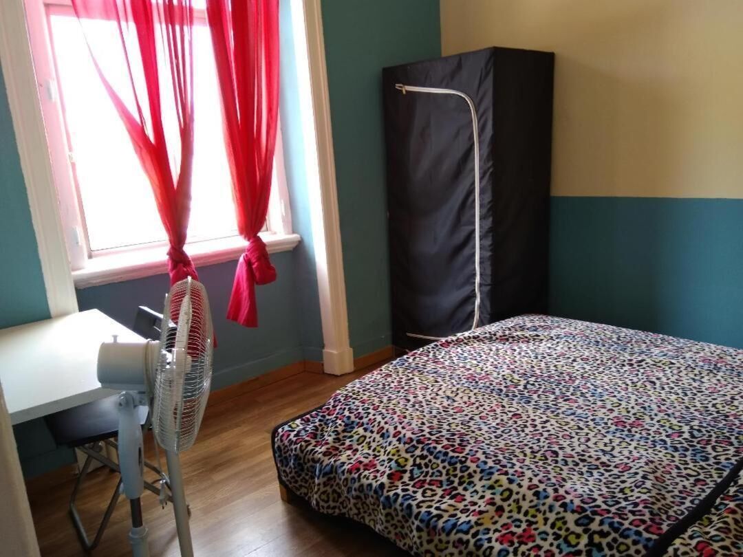 A bedroom with a bed, a desk, a chair, a standing fan, and a window with curtains.