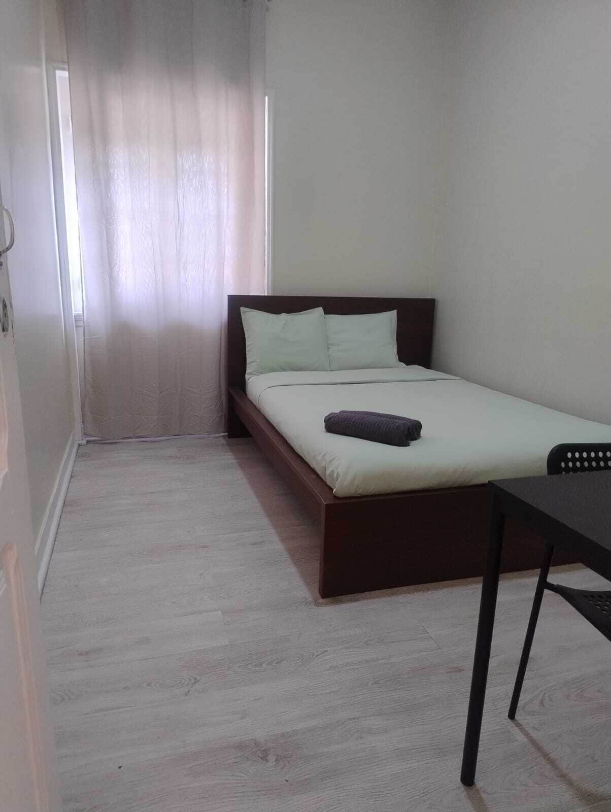 Een enkel bed met houten frame en matras, een klein tafeltje en een raam met gordijnen.